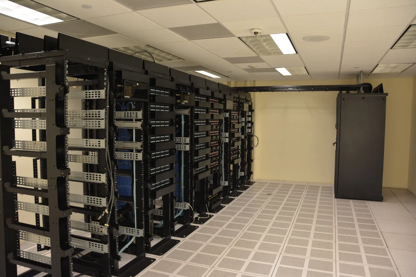 Interior of a data center with a row of open-frame server racks, organized network cabling, and a raised perforated floor under bright overhead lighting.