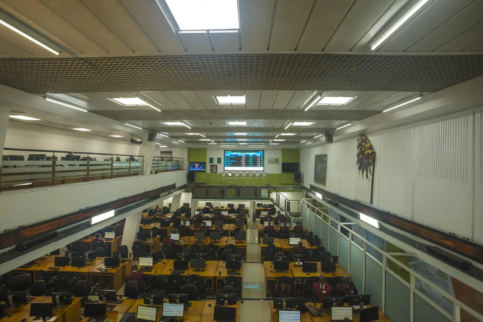 Wide view of a multi-level stock exchange trading floor with rows of dual-monitor workstations and a large market-data display board.