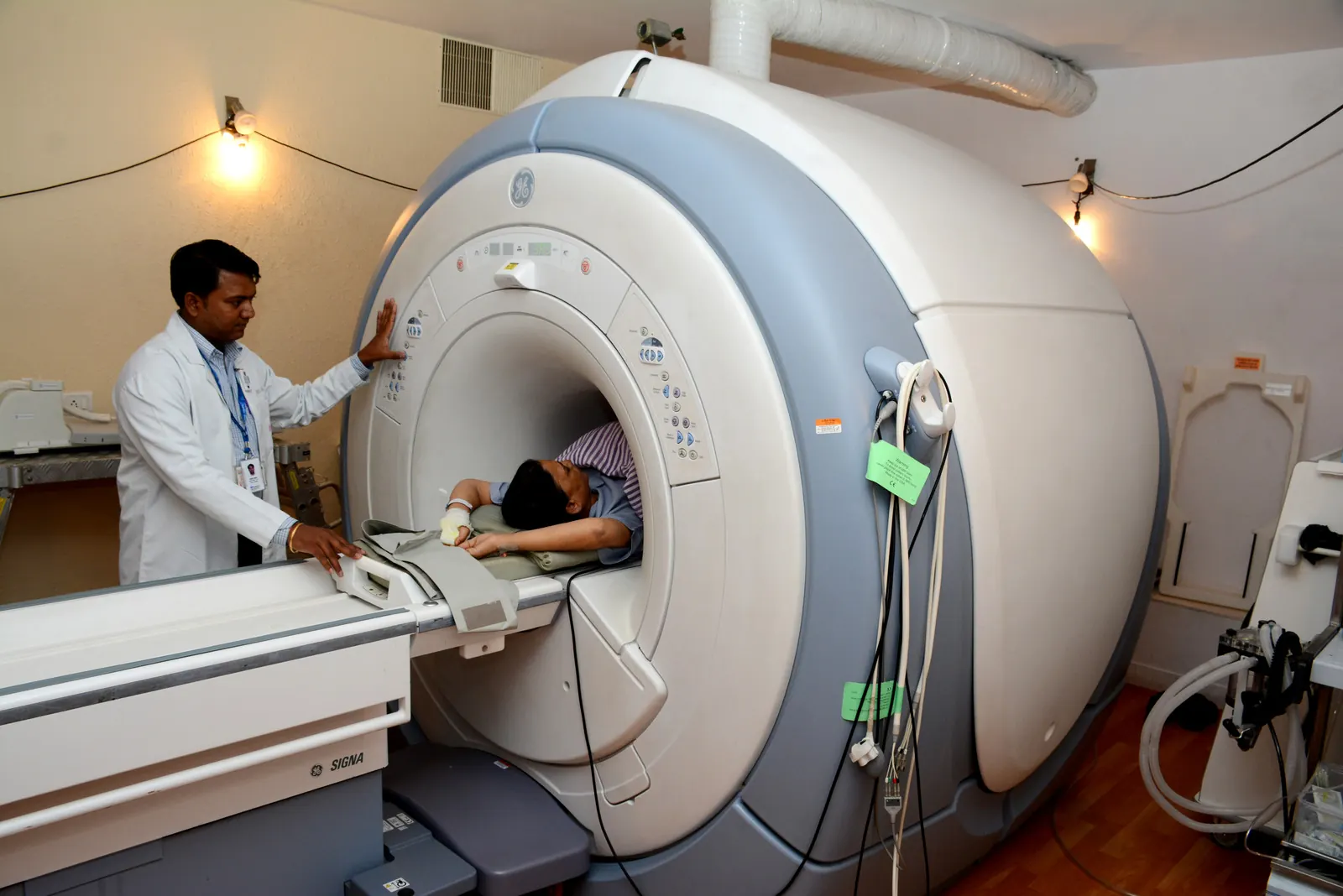 Radiologic technologist operates a GE Signa MRI scanner while a patient lies on the table in a hospital imaging suite.